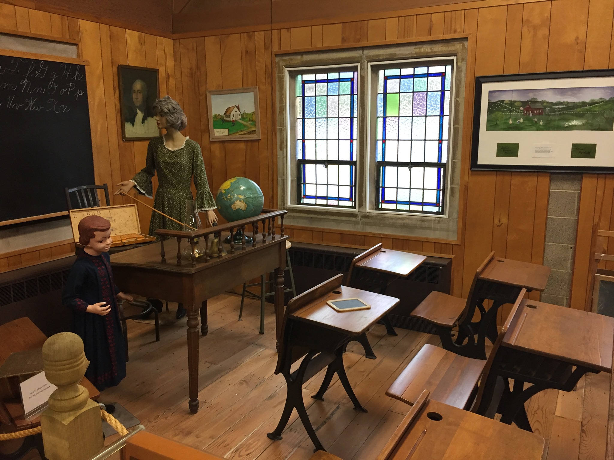 A museum exhibit of a 19th-century classroom featuring wooden desks, a mannequin teacher, and a chalkboard.